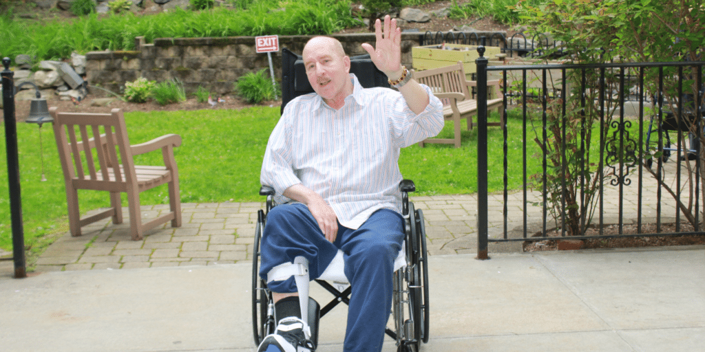 Photo of rehab patient in wheelchair waving at camera while sitting outside on patio