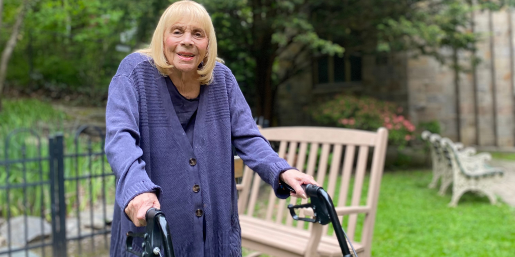 Woman smiling while standing with walker in outdoor garden, preparing to discharge home after rehabilitation.