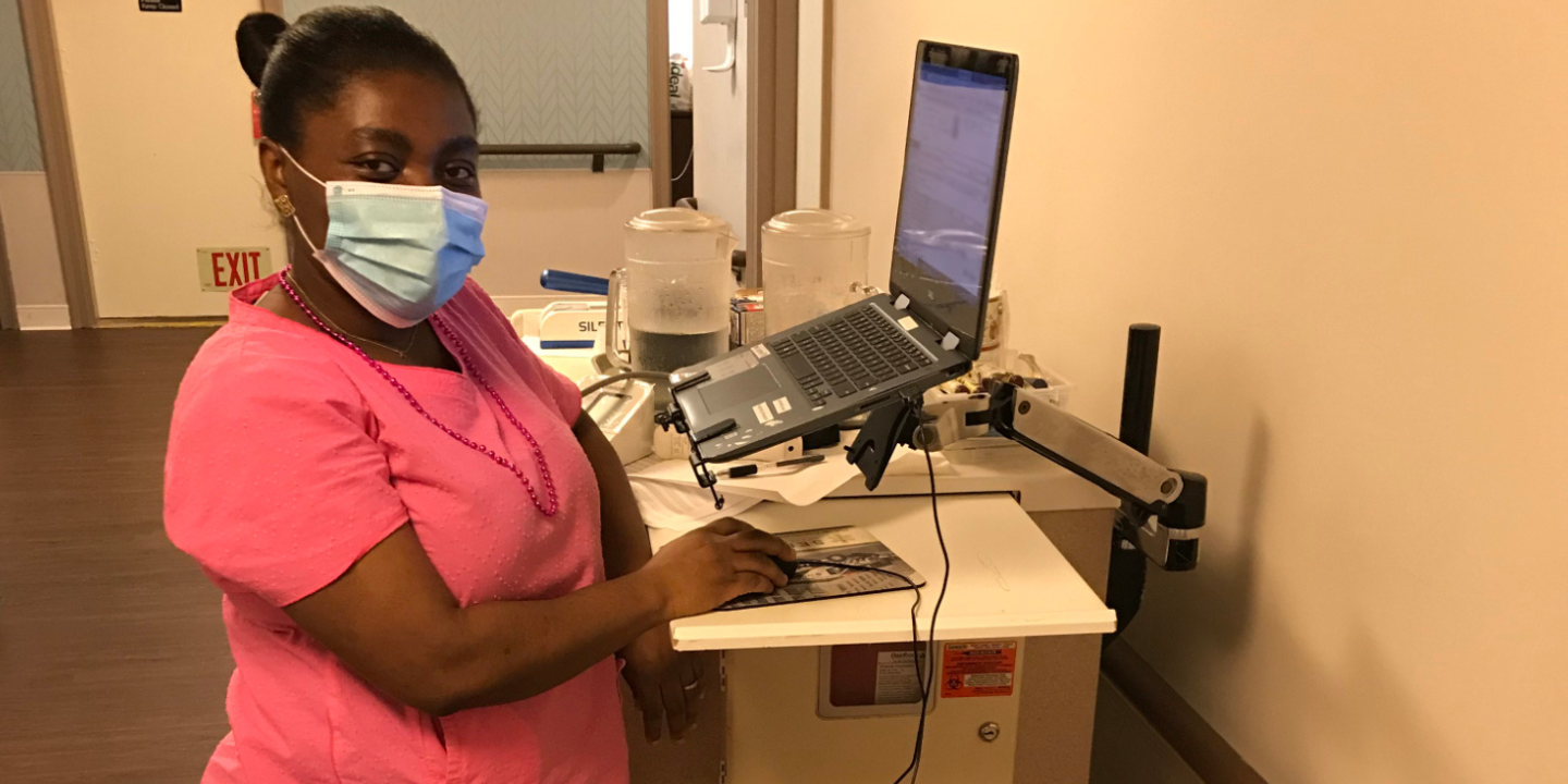 Photo of a nurse at standing cart with laptop wearing pink scrubs and a mask.
