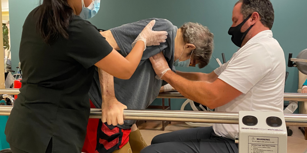 Photo of a short term rehab patient during an amputee rehabilitation session with his two therapists, they are helping him stand up from a wheelchair by supporting him with a grab bar for assistance.