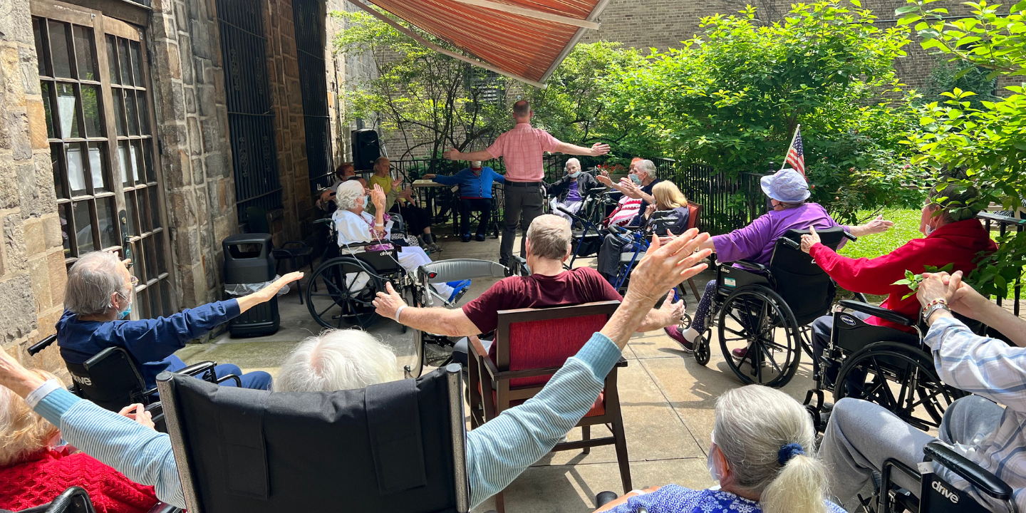 Photo of a group of skilled nursing facility residents in wheelchairs on outdoor patio enjoying morning stretch to work on mind body connection to start their day.