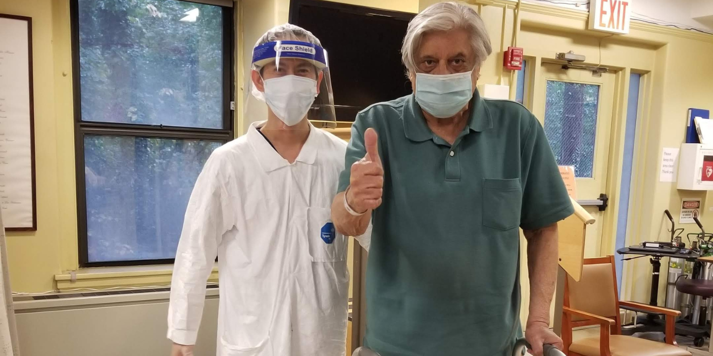 Photo of a senior man holding his thumb up in celebration during a therapy session, standing next to his physical therapist.