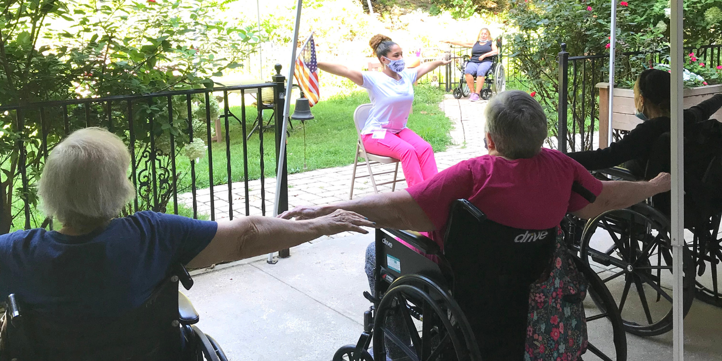 Therapeutic recreation services staff person sitting on a chair leading a chair yoga class with some skilled nursing facility residents on the patio following along to get morning stretch exercise.