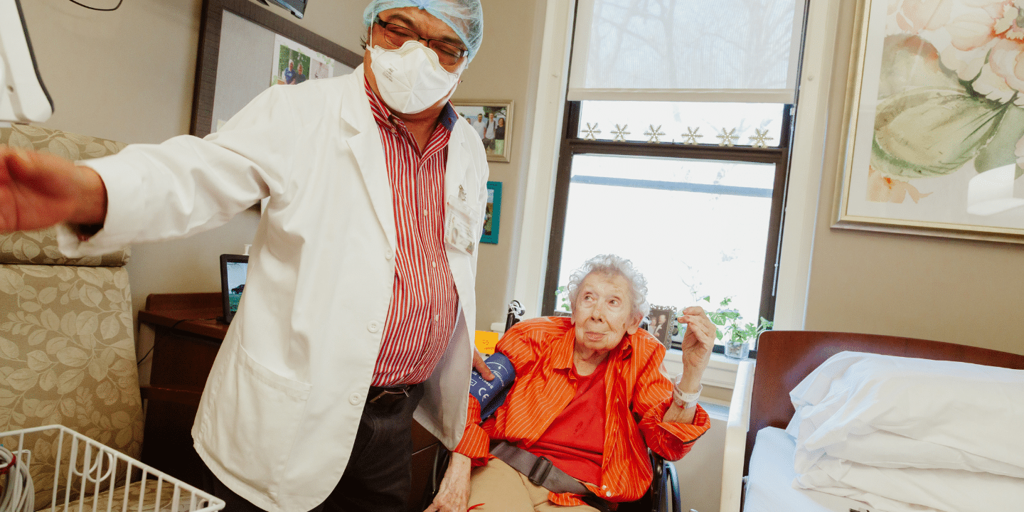 Photo of a senior woman sitting in a pretty patient room while her doctor takes her blood pressure, one of our patient needs.